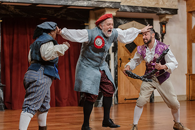 Dromio of Ephesus in boxing pose wearing blue vest and hat, blue striped kneepants with with stockings and work boots faces off against Antopholus of Ephesus holding a short black rope in his hands and wearing a fancy purple vest over white shirt and cream kneepants with white stockings and stylish shoes. The officer stands between them with outstretched hands planted on their foreheads; he's wearing a long vest coat with a red ribboned emplem, red cap, and maroon knee pants with black stockings.
